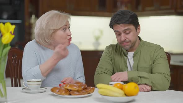 Disappointed Angry Senior Mother and Depressed Sad Adult Son Arguing Sitting at Table Indoors at