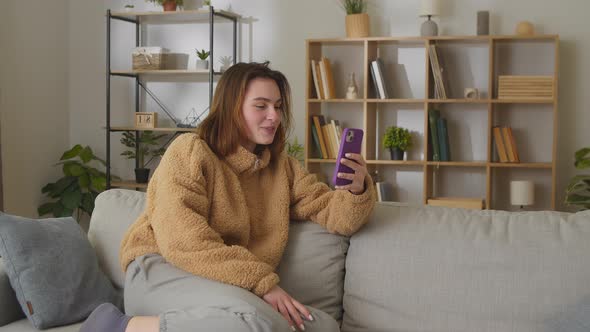 Pretty Happy Smiling Caucasian Young Woman Talking on Video on Phone While Sitting on Sofa in Room