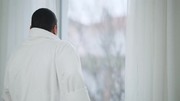 Back View of African American Young Man Putting on White Bathrobe Looking Out the Window in the