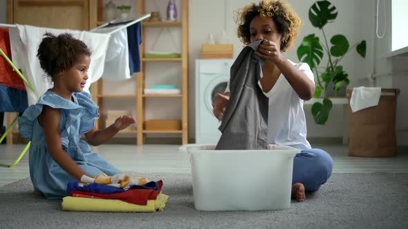 Young Mother and Cute Daughter Lay Out Clean Clothes and Sits on Floor in Laundry Room Spbd