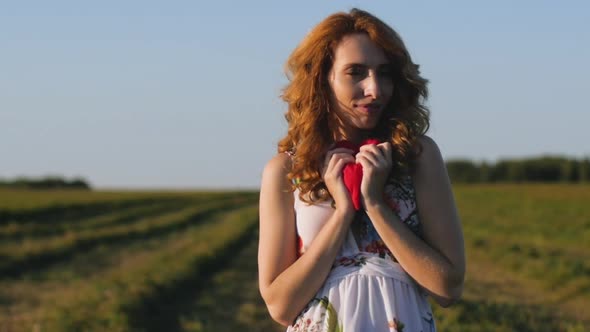 Redhaired Woman in a White Dress Holding a Soft Heart on Background of Field