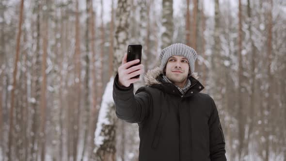 Caucasian Man in Winter Clothes Makes Selfie on Phone in Snowy Forest