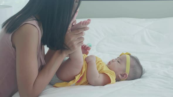 Young asian mother kiss foot of little baby girl with tender on bed in the bedroom.