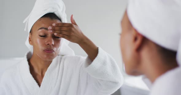African american woman in bathrobe applying face cream looking in the mirror at bathroom