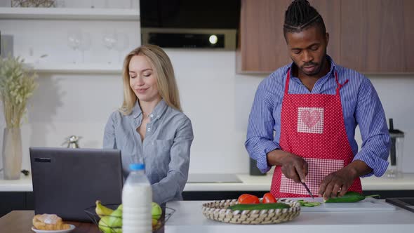 Handsome African American Husband in Apron Cutting Salad in Kitchen As Busy Caucasian Woman