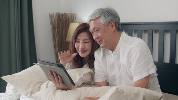 Asian senior couple using tablet video call talking with family grandchild kids while lying on bed.