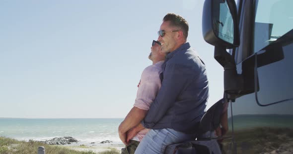 Happy caucasian gay male couple in sunglasses embracing, leaning on car on sunny day by the sea