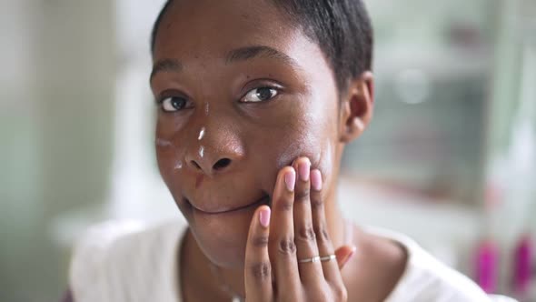 Headshot of African American Young Woman Applying Moisturizer on Face Looking at Camera