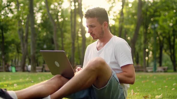 Young Man Sitting on the Grass in the Park Communicating By Video Call at Sunset