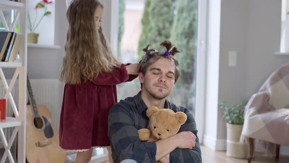 Concentrated Little Girl Making Ponytails on Hair of Joyful Father and Pulling Parent Ears
