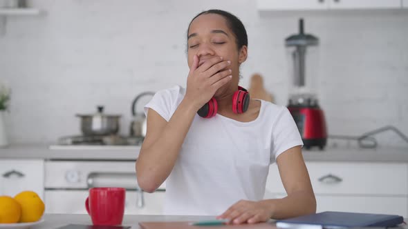 Exhausted Young Slim Woman Yawning and Putting Head on Laptop on Kitchen Table