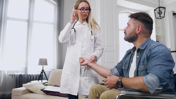 Professional Medical Assistant Checking Heart Rate to Balanced Young Bearded Man in Wheelchair