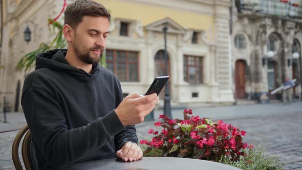 Hipster Guy with Mobile Smart Phone Digital Camera Talks While Sitting in Cafe