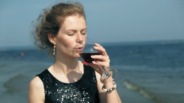 Young Blond Woman in a Beautiful Dress Enjoying a Glass of Pink Wine on the Beach Near the Sea