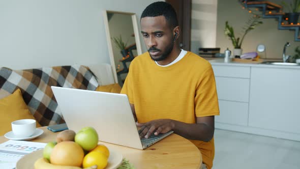 Portrait of Young Man Using Laptop Typing and Enjoying Music Through Wireless Earphones at Home in