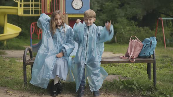 Cute Children Sitting on Bench on Playground and Putting on Raincoat Hoods