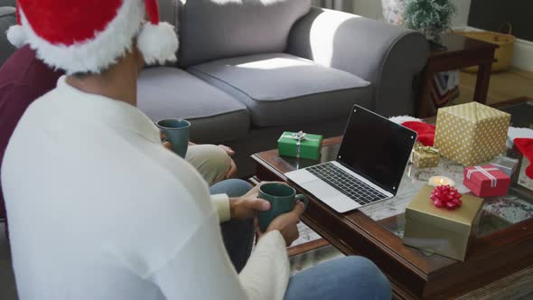 Father and son in santa hats having coffee while making laptop christmas video call at home