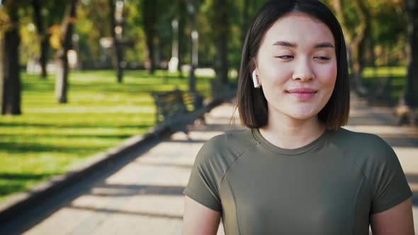 Adorable Asian Woman in Wireless Headphones Smiling to Camera in City Park Tracking Shot