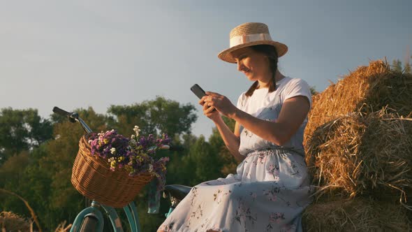 Woman is relaxing in meadow with smartphone in hands at sunset with retro bike next to her