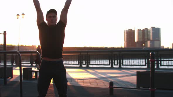 Young Sportive Man Squatting and Training His Hands with Dumbbells on Sunset