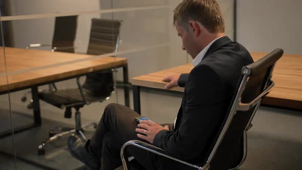 Young Handsome Businessman Looking at His Smartphone Sitting on Chair in the Office