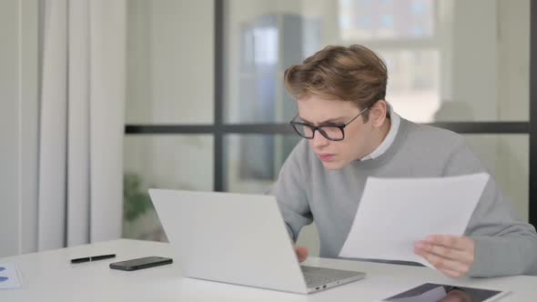 Young Man with Laptop Reacting to Loss While Reading Documents in Office