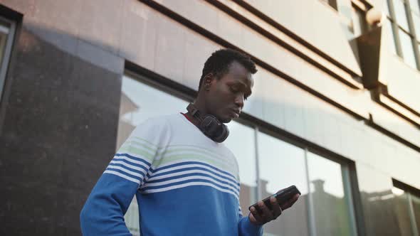 A Young Black Student in Stylish Clothes Sits and Talks on a Smartphone in the City