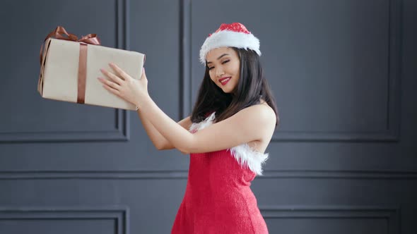 Smiling Asian Female Santa Claus Holding Big Carton Gift Box with Bow at Studio in Gray Background