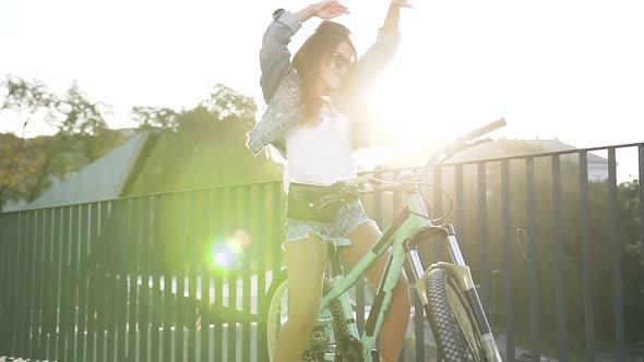 Girl in Stylish Clothing, Sunglasses with Long Hair Dancec in City while Standing with Bicycle