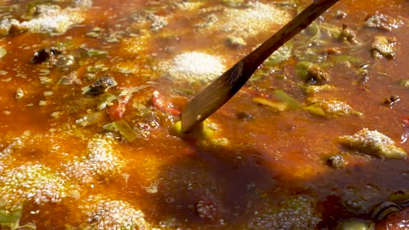 A close up shot of a wooden spoon mixing a soupy Spanish Paella while it is being cooked outside