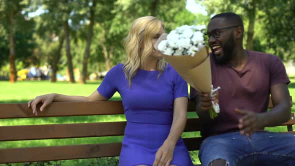 Black Man Presents Bouquet of Flowers to Sad Blond Woman Sitting in Park, Date