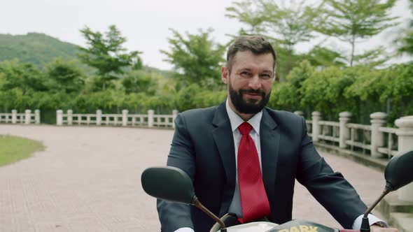 Portrait of a Senior Bearded Smiling Businessman Driving Up on a Motorcycle Along the Street