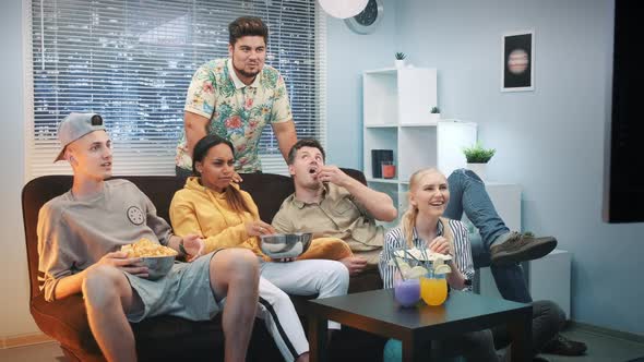 Excited Friends Relaxing on Sofa, Watching TV and Eating Snacks at Home