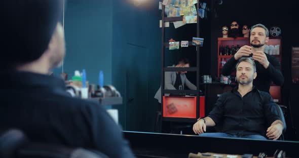 Adult Man Sits in a Stylist's Chair Opposite the Mirror