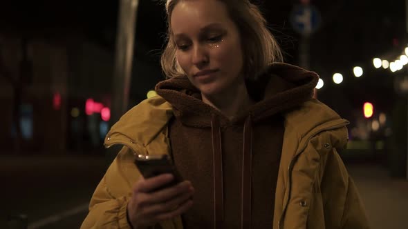 Short Haired Girl Walks Through the Evening City Smiling Stops and Texting in the Smartphone