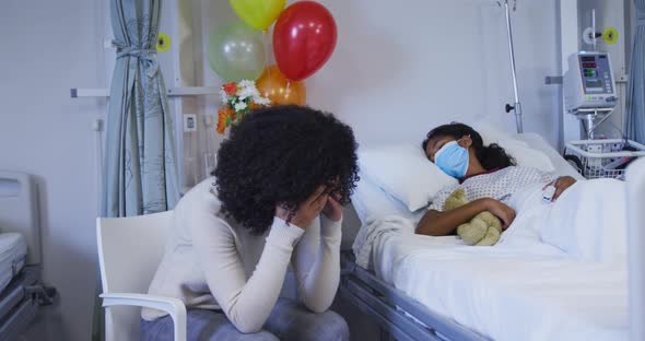 Stressed african american mother sitting besides her her daughter lying on bed at hospital