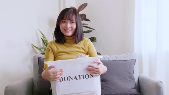 Portrait of young Asian woman looking at camera holding box with clothes for donation. Concept campa