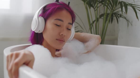 Young Asian Lady Lying in Bathtub and Listening to Music with Headphones