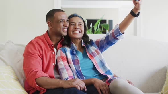 Happy biracial couple sitting on sofa, embracing, using smartphone and laughing