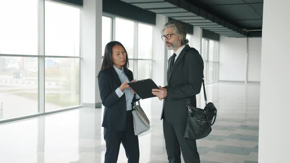 Asian Businesswoman Talking to Partner Studying Papers Standing Indoors in Modern Glass Walls