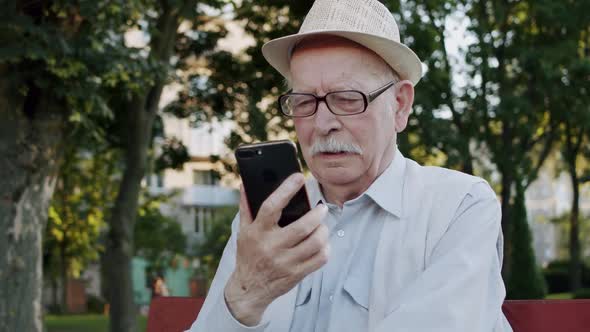 Thoughtful Senior Man with Glasses Chatting Seriously on Phone's Camera in Park