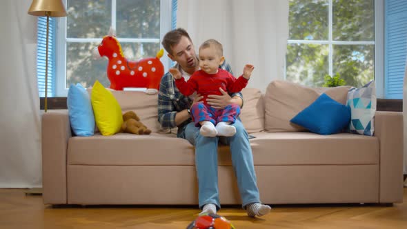 Young Man Playing with Toddler Son Sitting Together on Couch at Home