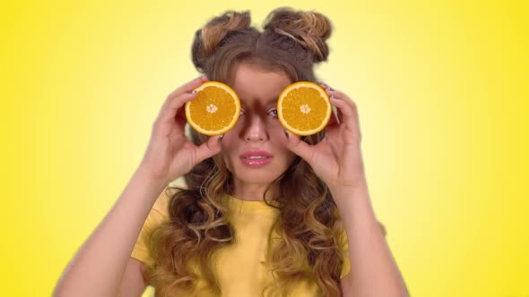 Attractive Girl in a Yellow Vest Closes Her Eyes with Oranges and Smiles Looking Into the Camera