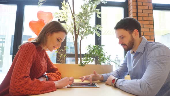 Happy Young Couple a Guy and a Girl Are Sitting at a Table in a Cafe and Using a Tablet