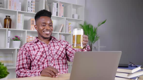 African man working from home taking vdo call on laptop with colleague and drinking beer 