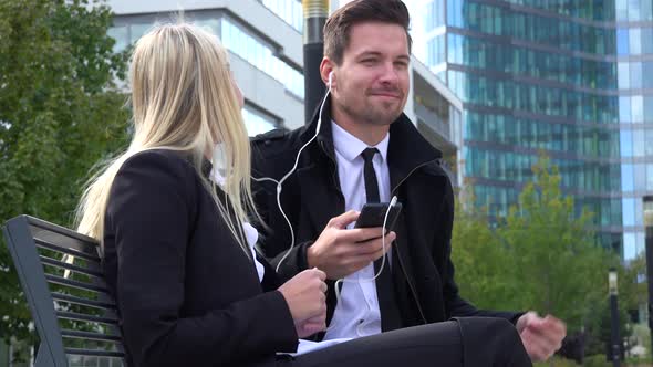 A Businessman and a Businesswoman Sit on a Bench and Listen To Music on a Smartphone in Urban Area