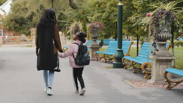 Back View African Woman Mother Holds Little Afro American Girl By Hand Going in City Park Back to