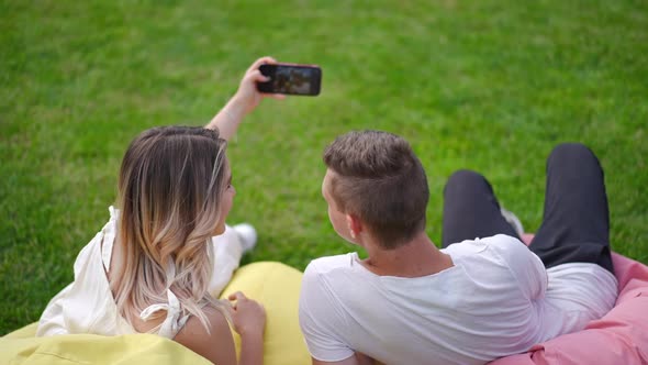 Back View Loving Positive Couple Taking Selfie on Smartphone Sitting on Bag Chairs on Green Meadow