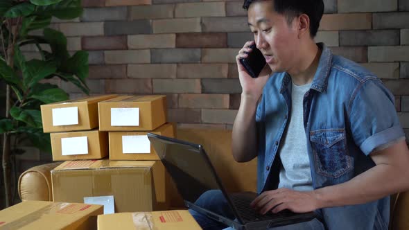 Asian Male Business Entrepreneur Using Laptop and Phone with Packs of Boxes at Home