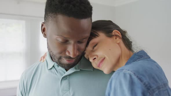 Happy diverse couple wearing jacket and shirt and embracing in living room